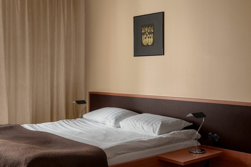 A neatly made bed with white sheets and two pillows, flanked by two modern black lamps, with beige curtains and a framed artwork above the headboard.