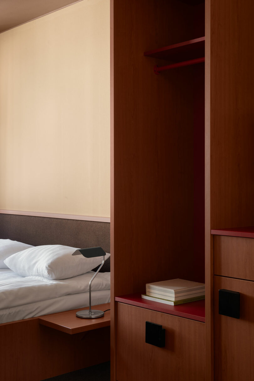 A minimalist hotel room with a neatly made bed, a small bedside table with a lamp, and an open wooden wardrobe with shelves containing stacked books.