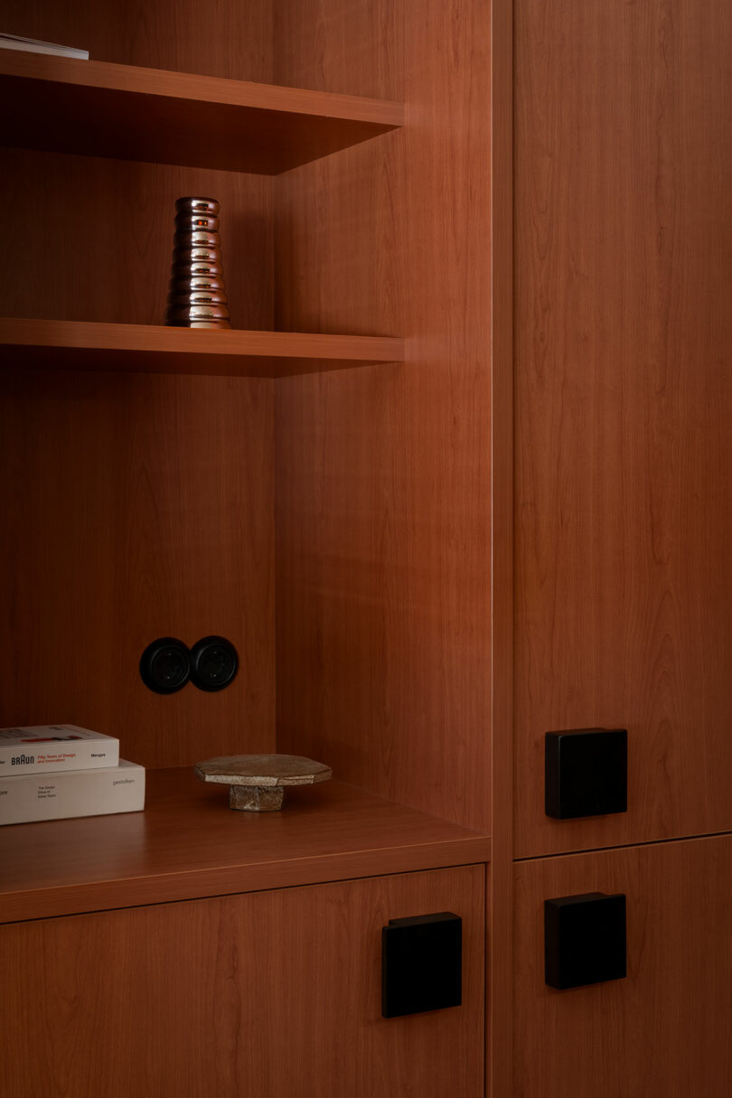 A wooden cabinet with black square handles, an open shelf holding a stacked metallic object, and a countertop with books and a decorative bowl.