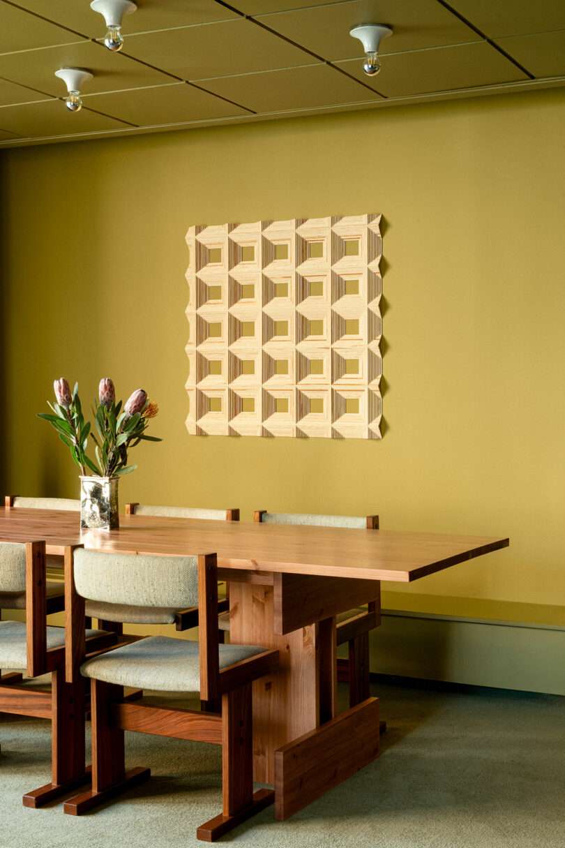 A wooden dining table with four chairs is set against a mustard yellow wall, decorated with a geometric wooden art piece; a vase with flowers sits on the table.