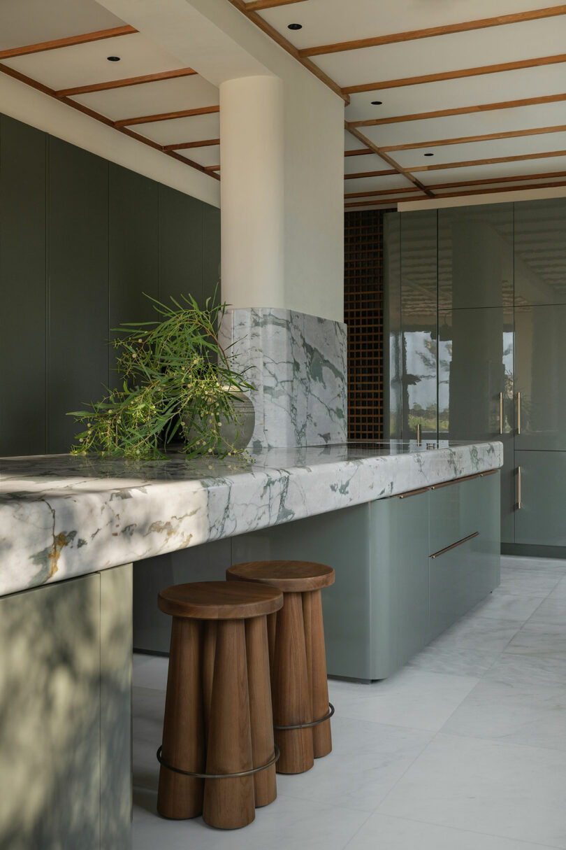 Modern kitchen with green cabinets, marble countertops and backsplash, wooden stools, and a potted plant on the counter. Natural light enters from the left.