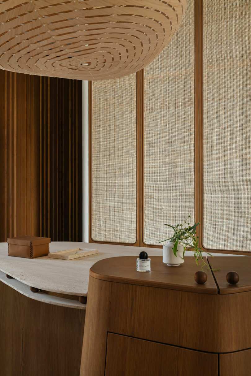 A modern interior with wooden furniture, woven paneling, a large wooden pendant light, a tray, a small potted plant, and a bottle on a curved wooden surface.
