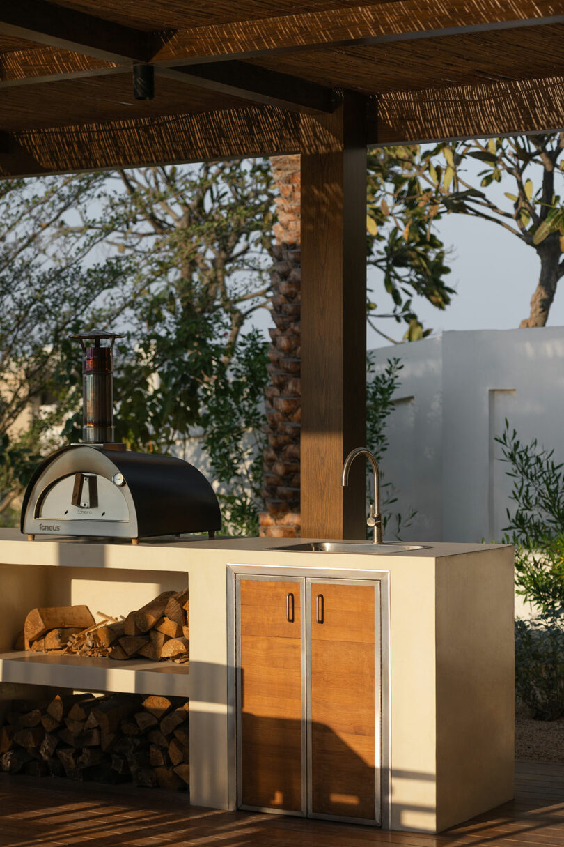 Outdoor kitchen with a built-in wood-fired oven, stacked firewood, countertop sink, and wooden cabinet doors under a pergola, surrounded by greenery.