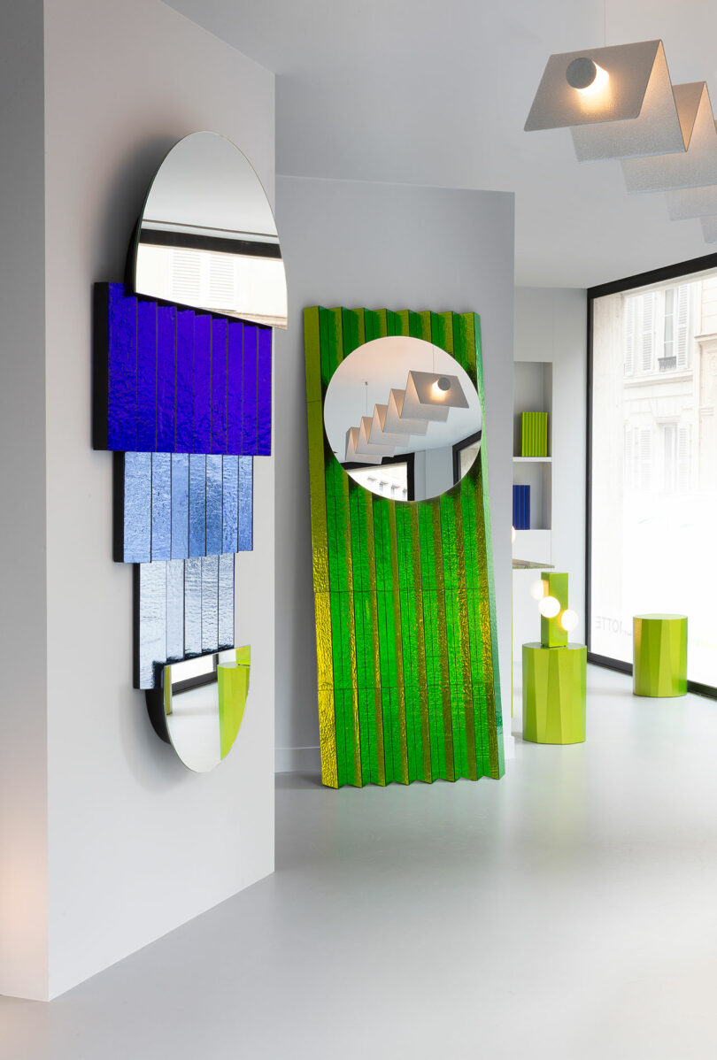 A modern gallery space featuring Colorscape by Victoria Wilmotte, with geometric mirrors and textured colored glass panels in blue, green, and yellow on white walls and floor, complemented by large windows in the background.