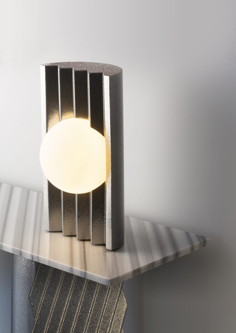 A round light is partially enclosed by a textured metallic frame with vertical slats, sitting on a geometric, striped surface against a plain background.