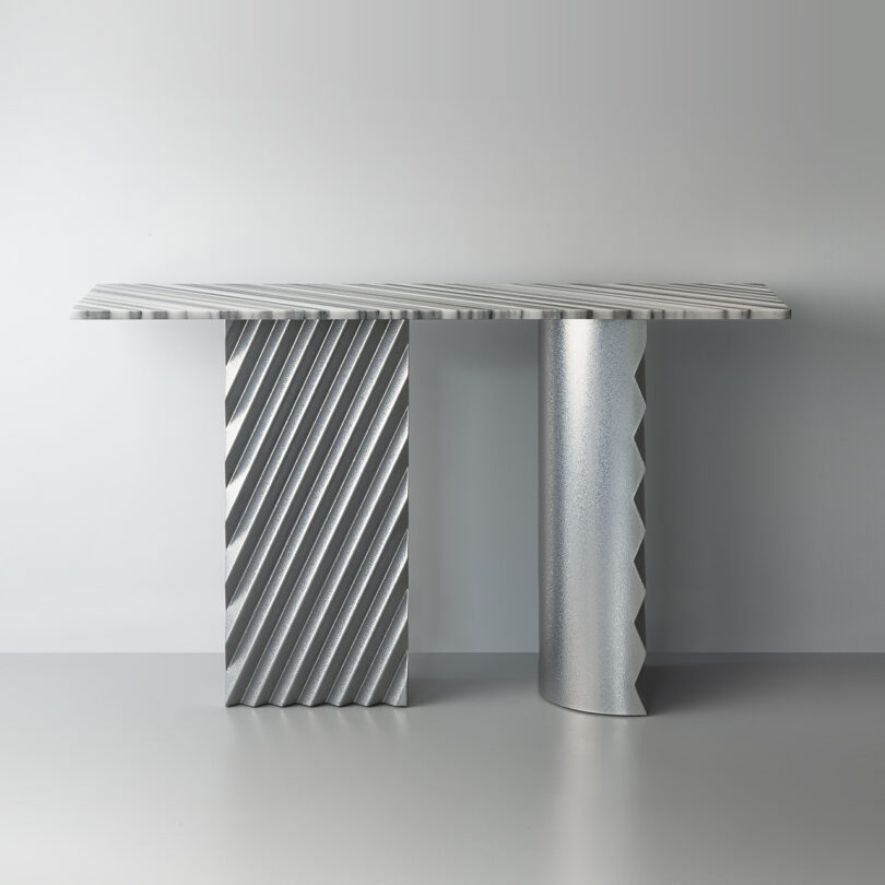 A modern metallic table with a flat rectangular top, supported by one rectangular and one cylindrical leg, both featuring diagonal and zigzagged textured patterns.