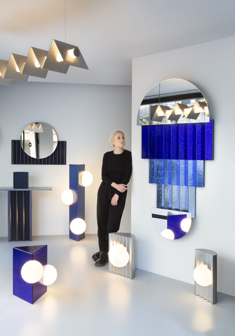 A person in black stands beside a large mirror with blue geometric accents, surrounded by modern light fixtures and reflective surfaces in a minimalist room.