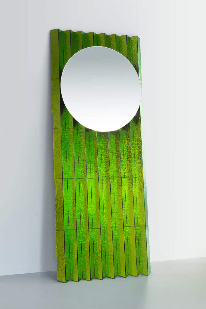 A tall, rectangular, textured green panel with vertical ridges stands against a wall, featuring a large round mirror near the top.