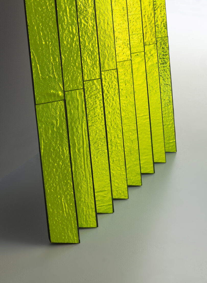 Nine vertical panels of textured yellow-green glass are arranged in a row, partially overlapping each other, against a light background.