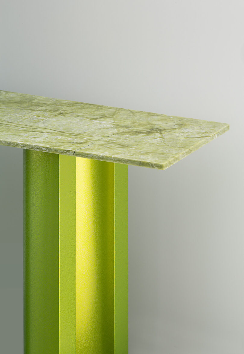 A table with a thick, green marble top and a metallic green rectangular leg against a plain light background.