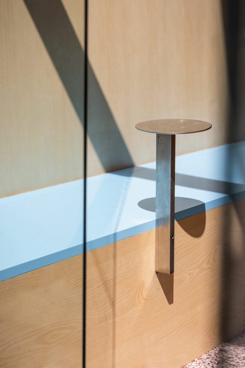 A small round table with a metal support attached to a light wood bench, with shadows cast by sunlight through glass.