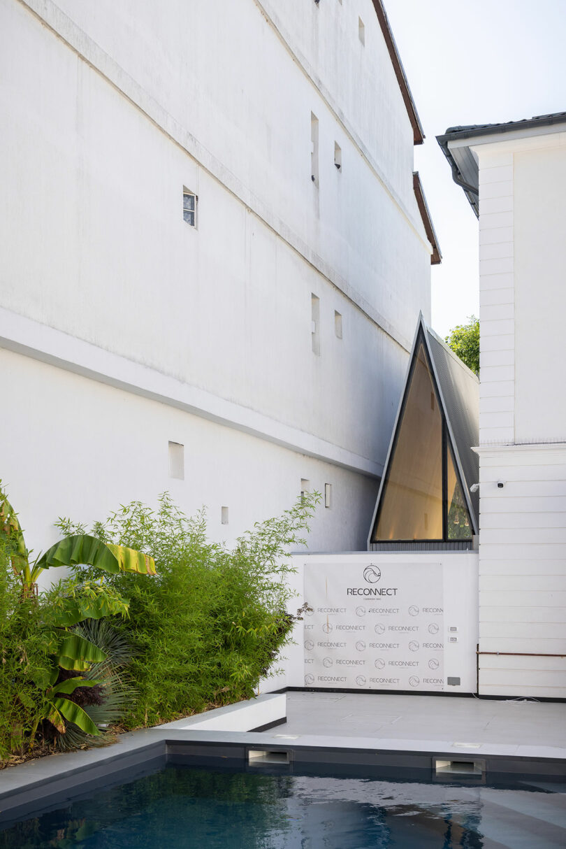 A small, modern pool is adjacent to a white building with a triangular glass structure and a backdrop displaying the 