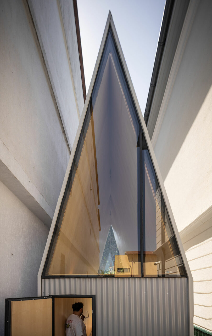 Tall, narrow building with a steep triangular glass facade situated between two white walls; a person stands at the open entrance below.