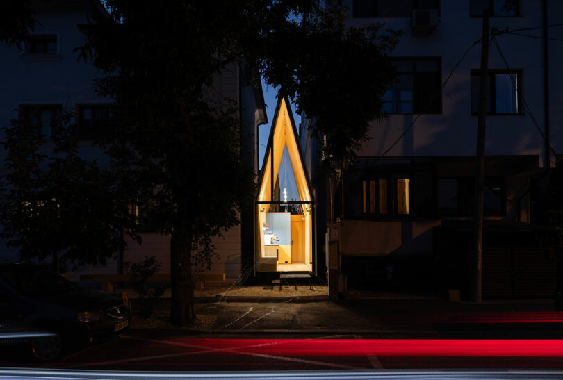 A narrow, modern house with a steep A-frame roof is illuminated at night between two larger buildings, with car light trails visible in the foreground.