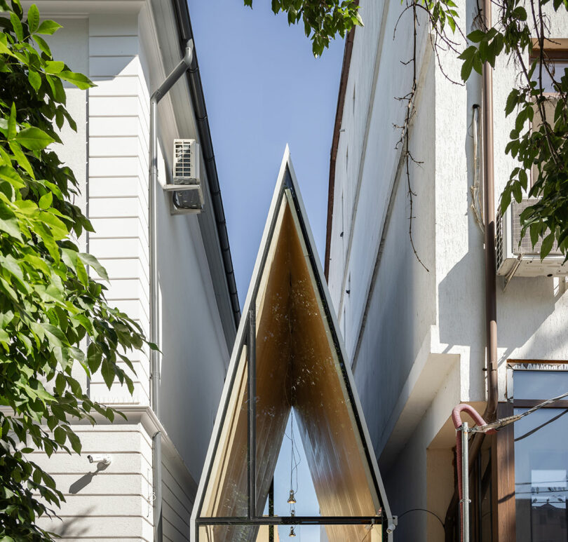 A narrow, triangular glass structure is positioned between two white buildings with exterior air conditioning units and leafy branches partially framing the view.