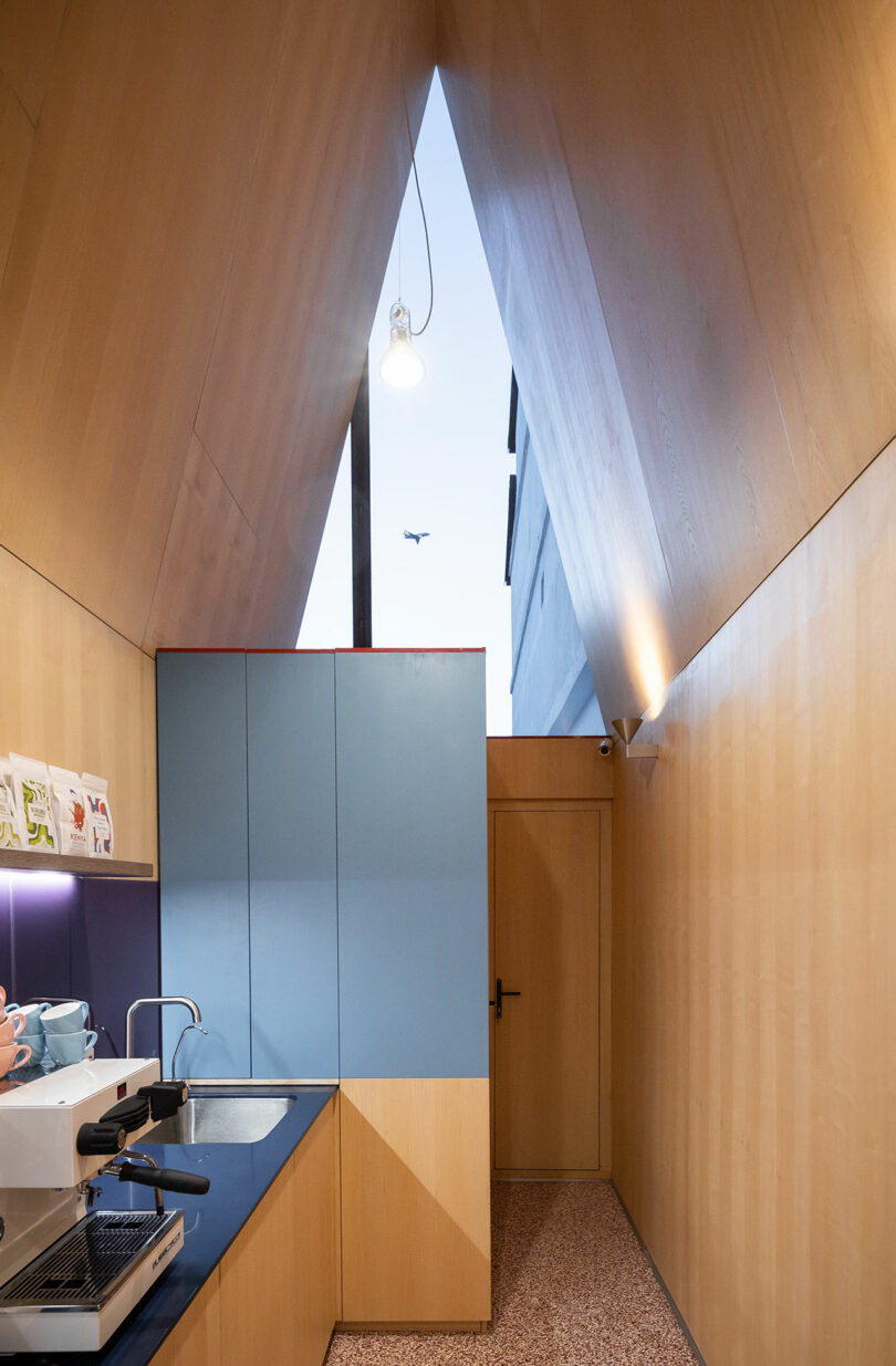 A small modern kitchen with wooden walls, blue cabinets, a coffee machine, and a sharply peaked ceiling with a window showing an airplane in the sky.