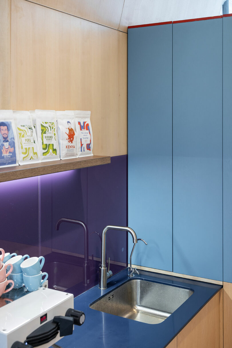 A modern kitchen corner with a blue countertop, stainless steel sink, coffee machine, stacked cups, and bags of coffee on a wooden shelf above a purple backsplash.