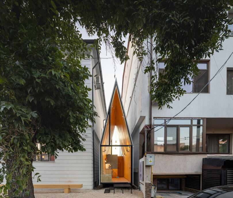 A narrow, modern, A-frame building with a glass facade is situated between two larger buildings, framed by trees in the foreground.