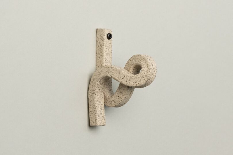 A beige, speckled ceramic hook with a looped design is mounted on a plain, light gray wall