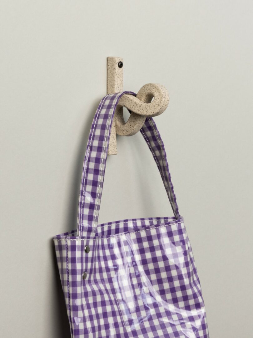 A purple and white checkered bag hangs from a beige, textured hook on a light gray wall