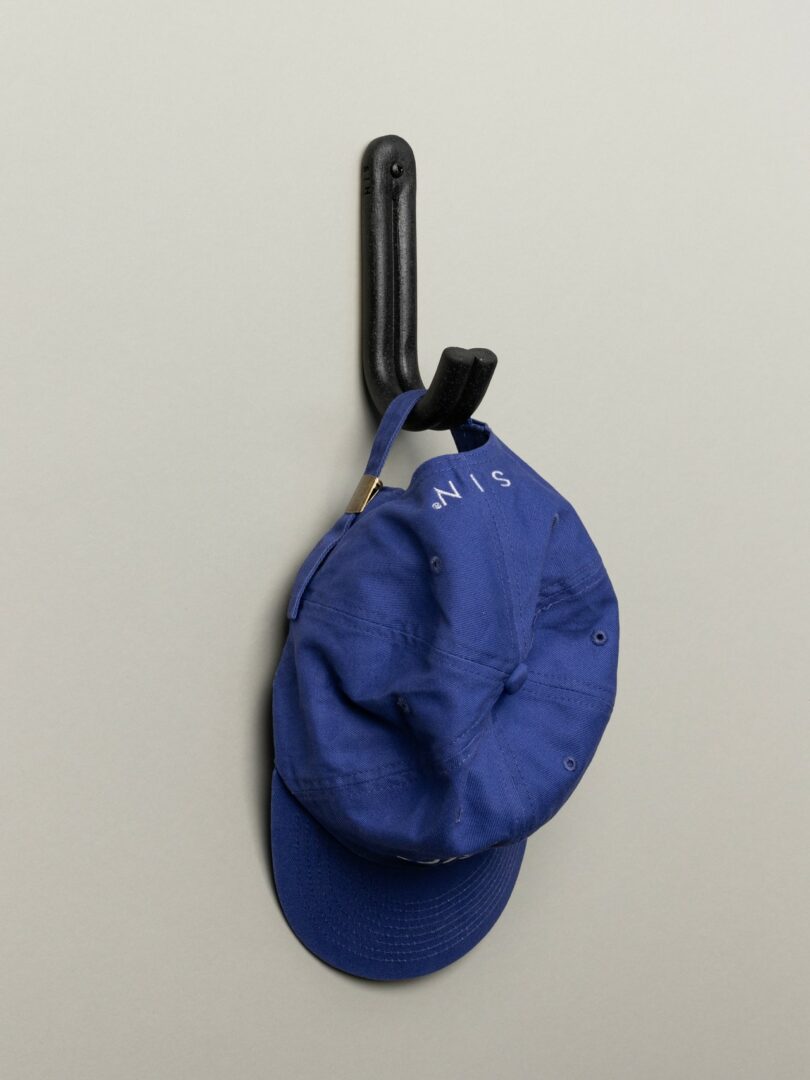 A blue baseball cap hanging on a black wall hook against a plain beige background