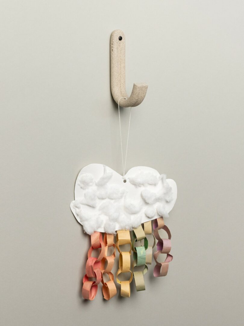 A handmade paper cloud with cotton and colorful paper chain rain hangs from a hook on a plain wall