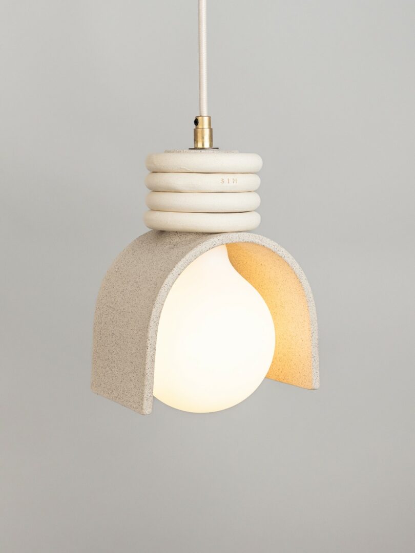 A pendant light with a round white bulb covered by a textured, arch-shaped beige lampshade hangs from a white cord against a plain gray background
