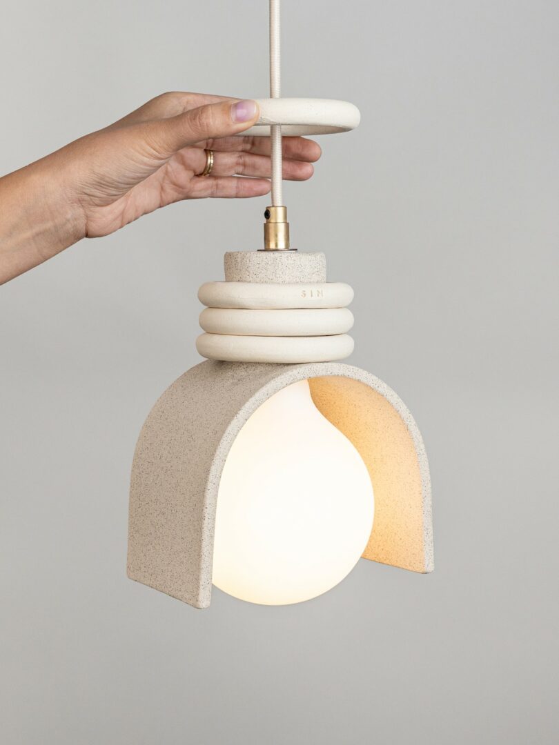 A hand holds a pendant light with a textured, off-white ceramic fixture and a round, glowing bulb, set against a plain gray background