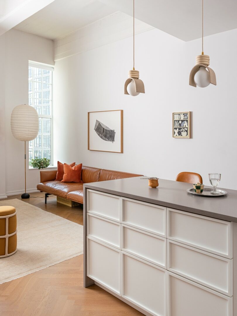 A modern, minimalist living room and kitchen with a brown leather sofa, white island, pendant lights, and large windows allowing natural light