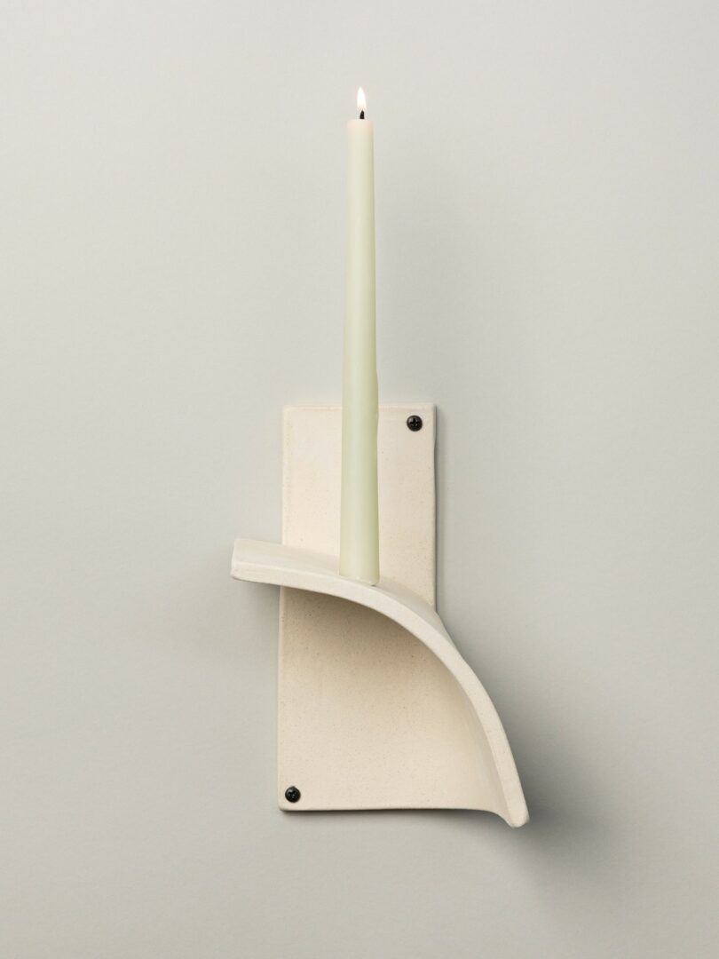 A white, minimalist ceramic wall sconce holds a lit tapered candle vertically; the sconce features a curved ledge design and two visible screws