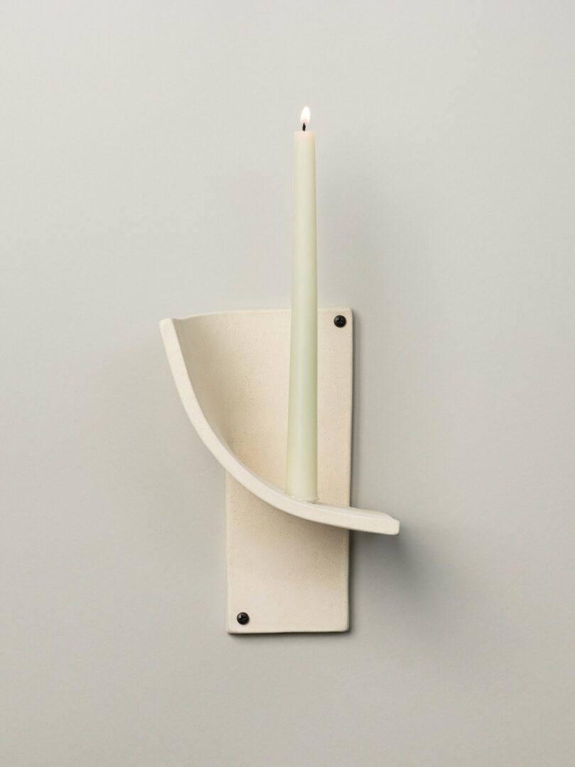 A white ceramic wall-mounted candle holder with a single lit cream-colored taper candle against a plain light background