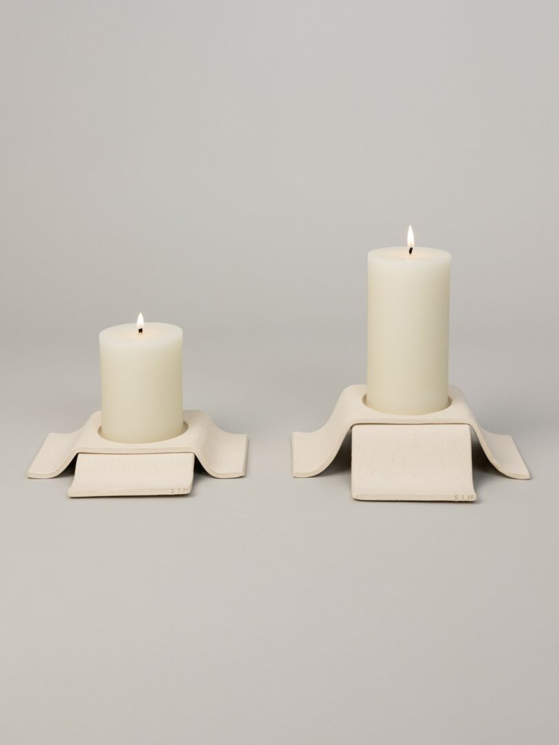 Two lit white pillar candles placed on modern, rectangular cream-colored ceramic holders with curved edges, set against a plain light gray background