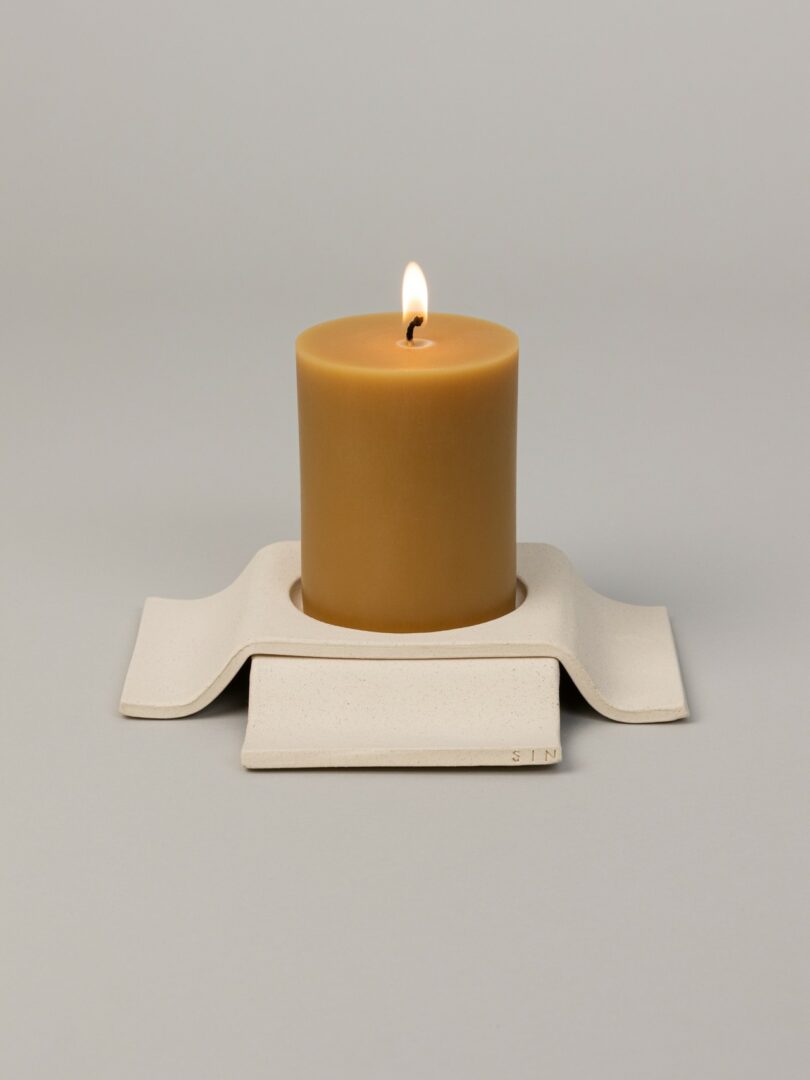 A lit brown pillar candle sits on a sculptural, wavy white ceramic holder against a plain light gray background