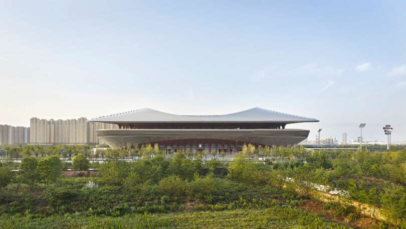 A modern stadium with a curved roof stands amid greenery, with high-rise buildings visible in the background under a clear sky.