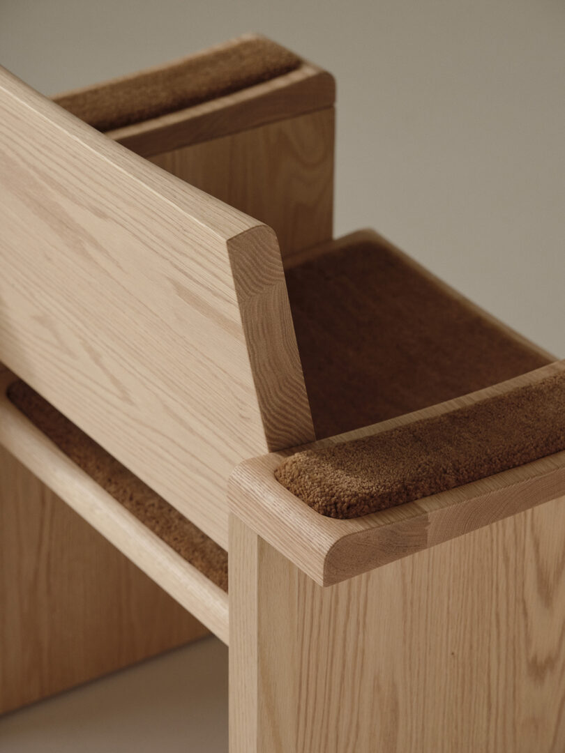 A close-up view of a wooden chair with a light wood finish, featuring angled backrest and armrests, and brown padded seat and armrest cushions.