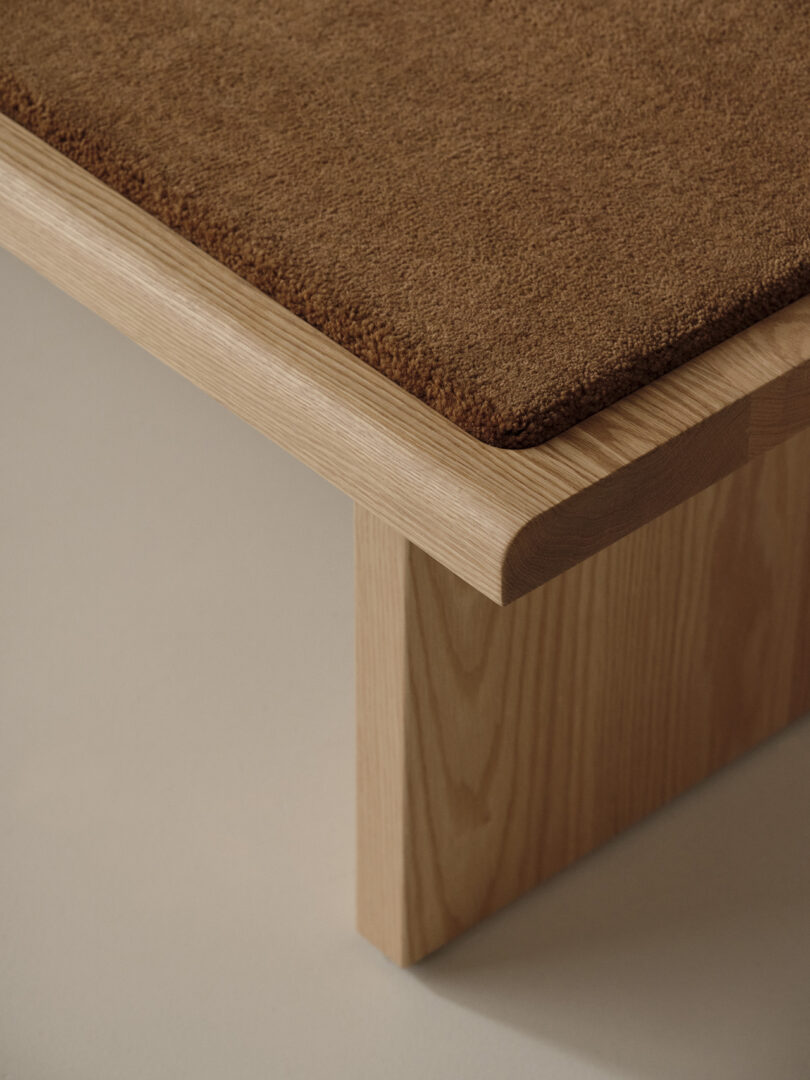 A close-up of the corner of a wooden furniture piece, possibly a bench or table, with a textured brown cushion or mat on top.