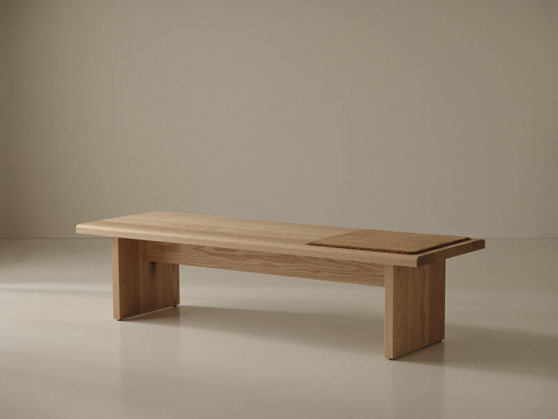 A minimalist wooden bench with a rectangular seat and sturdy legs, featuring a section with a woven or textured surface on one end, set against a plain background.