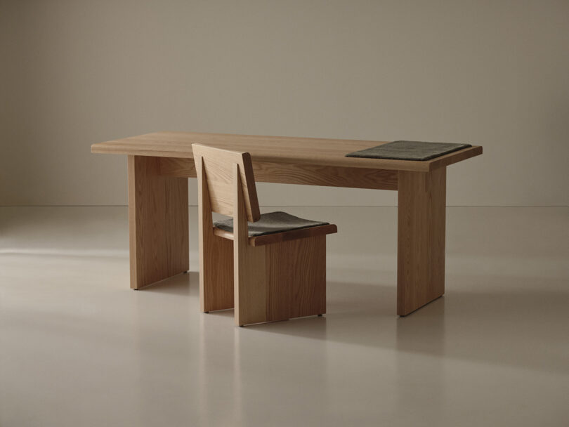 A wooden desk and matching chair with straight lines are placed in a plain room. Two rectangular gray mats sit on the desk and chair seat.