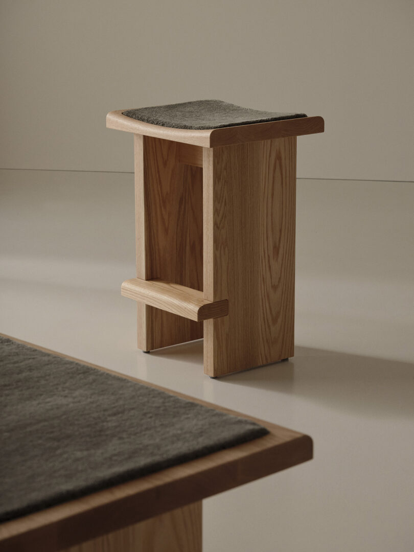 A minimalist wooden stool with a flat gray cushion sits on a smooth, light-colored floor. Part of a matching bench is visible in the foreground.