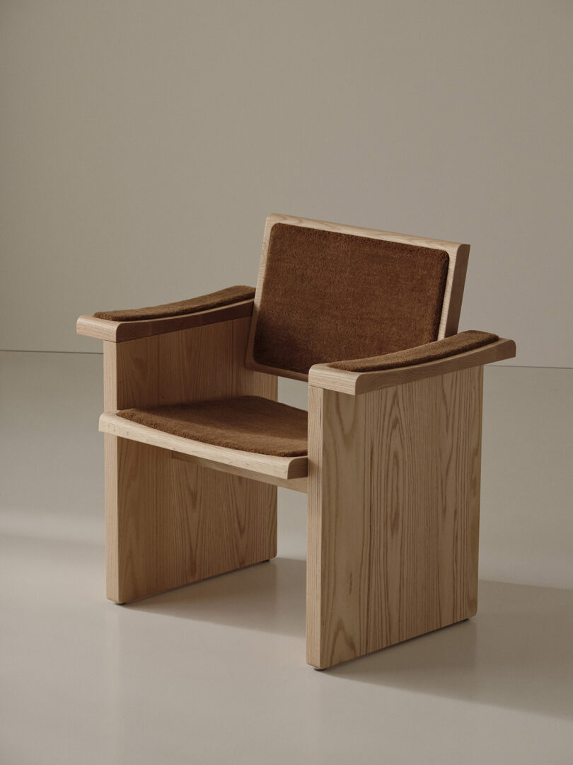 A wooden armchair with a minimalist design, featuring a rectangular frame and flat armrests, with brown cushioned seat, back, and arm pads.