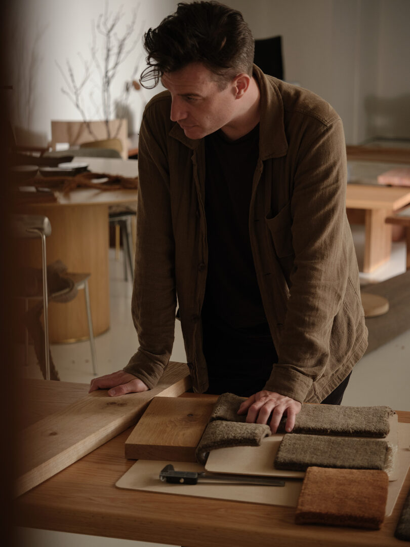 A man stands at a wooden table, looking down at samples of wood and fabric, with a measuring tool nearby in a workspace.