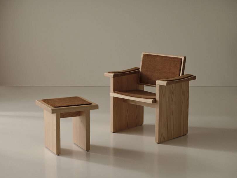 A wooden chair with a brown seat and backrest pad is positioned next to a matching wooden stool in a minimalist room with a neutral background.