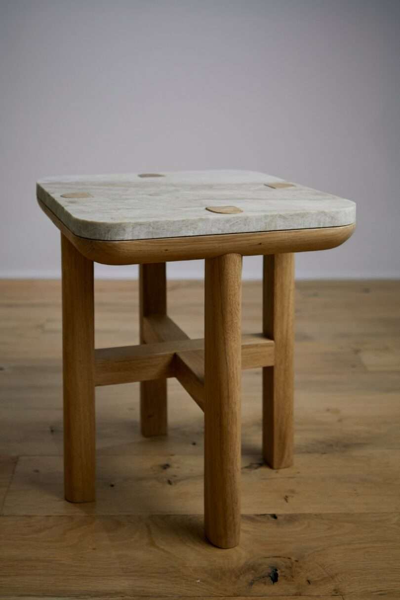 A wooden stool with four legs and a light-colored, square seat is placed on a wooden floor against a plain gray wall