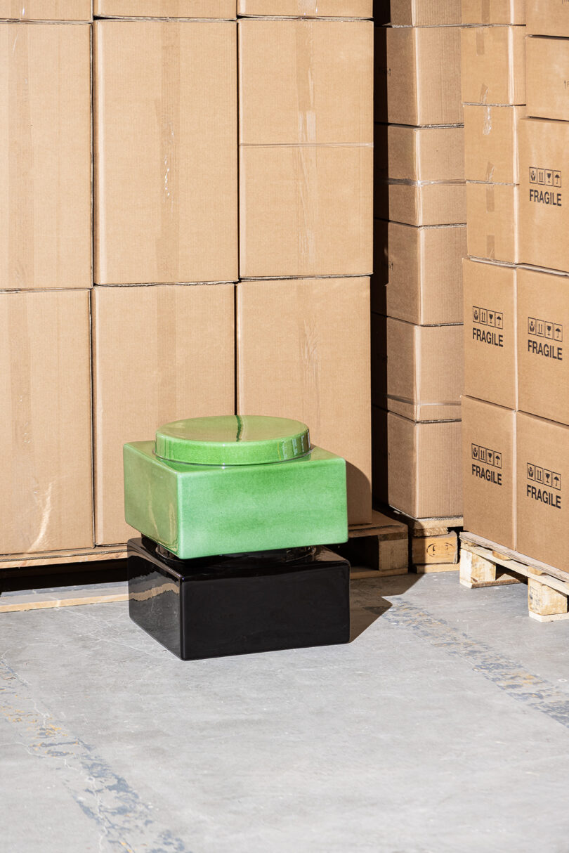 A green, square-shaped ceramic object stacked on a black rectangular base sits on a concrete floor in front of cardboard boxes labeled 