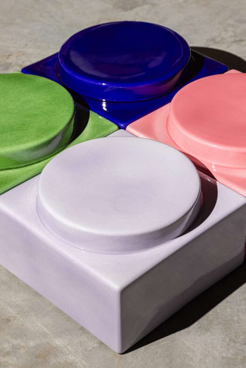 Four glossy, round ceramic shapes in blue, green, pink, and lavender are embedded in a square lavender base, placed on a concrete surface.
