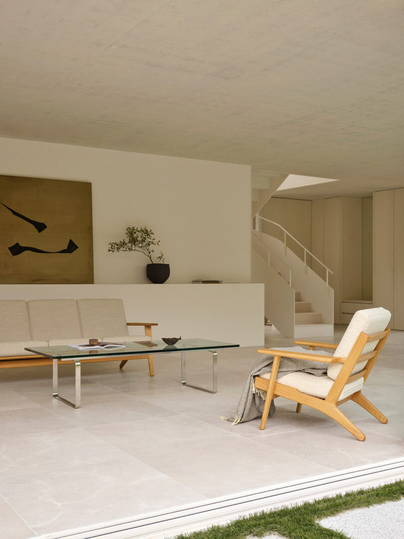 Minimalist living room with light-colored furniture, a glass coffee table, abstract wall art, a potted plant, and stairs in the background. The room opens to a grassy outdoor area.