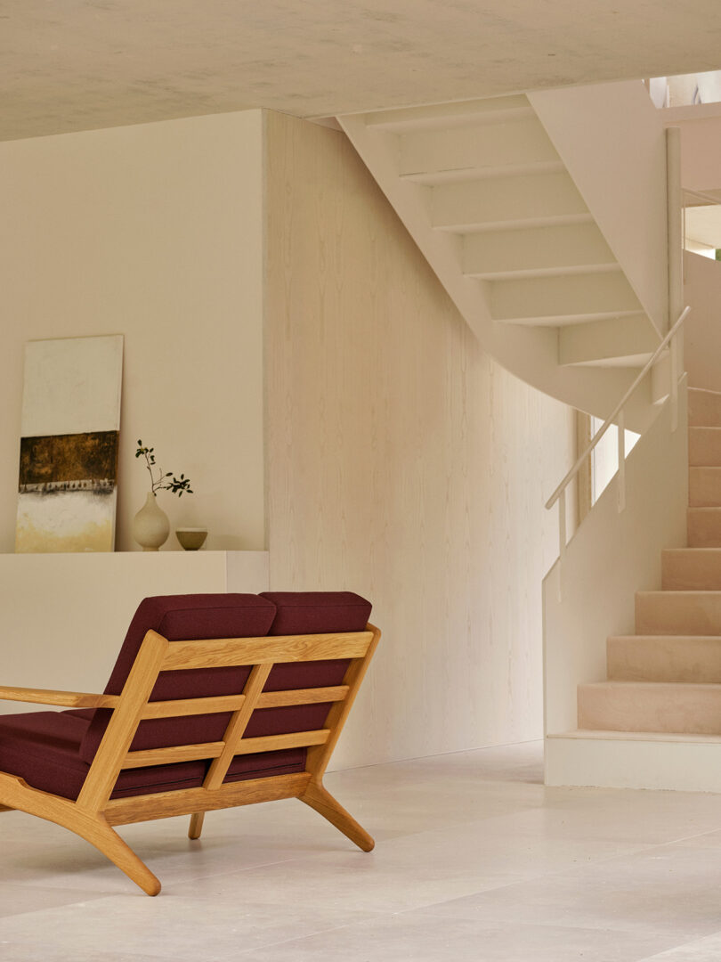 A modern interior featuring a wooden chair with burgundy cushions, a minimalist side table with decor, and a white staircase with a curved underside.