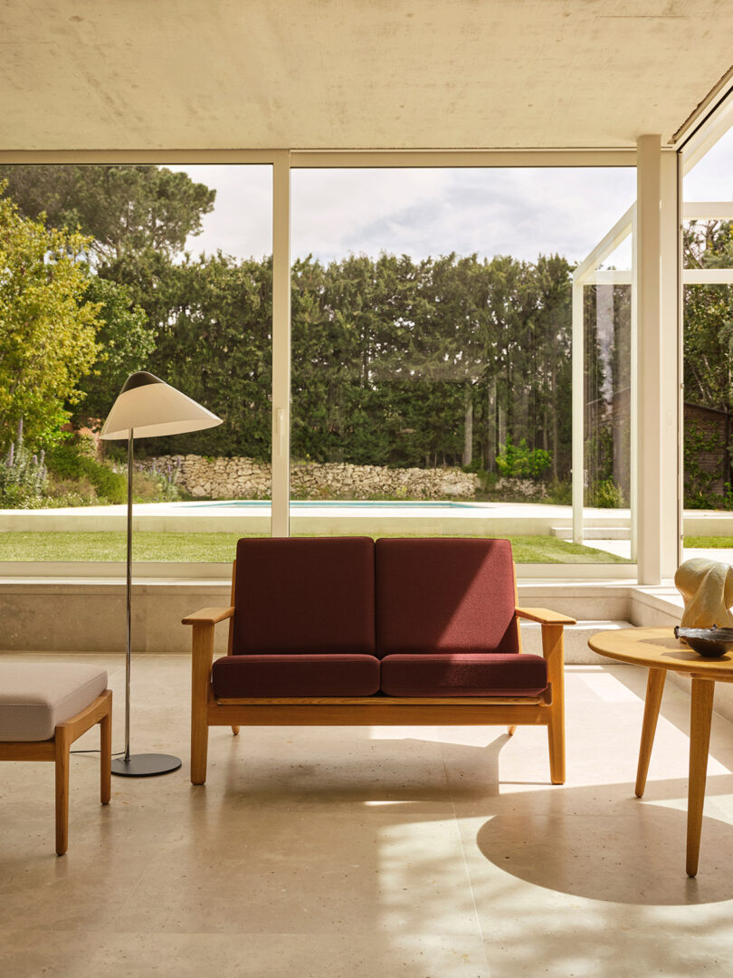 A minimalist living room with a maroon loveseat, round wooden side table, and floor lamp, featuring large windows overlooking a green garden.