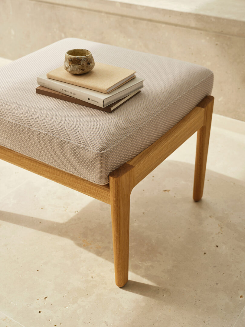 A light wood bench with a textured beige cushion holds three stacked books and a small ceramic cup, set on a beige stone floor.