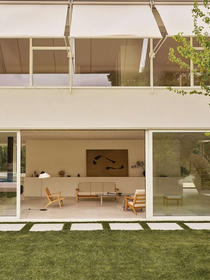 Modern living room with minimalist furniture and artwork, seen through large glass sliding doors; exterior shows green grass and stone pavers.